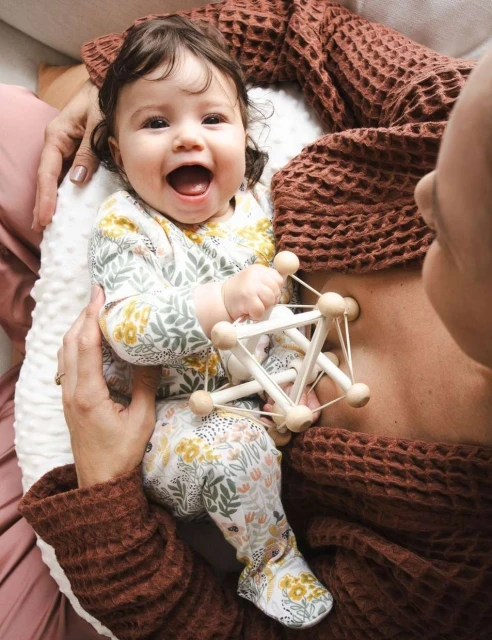 Happy baby in floral print outfit lies next to a woman wearing a brown waffle-knit top while holding a wooden geometric toy.