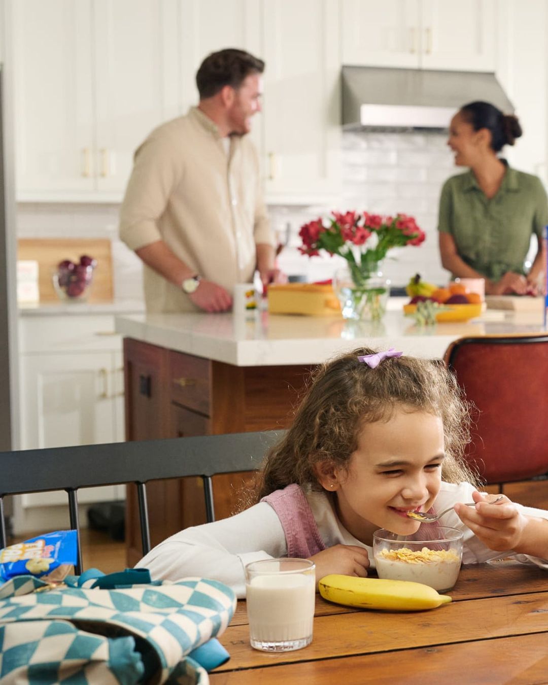 A little girl eats cereal at a wooden table indoors, with her parents standing by the kitchen island in the background.