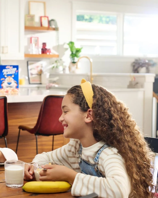 A cheerful girl with curly hair enjoys a breakfast of cereal with milk and a banana in a brightly lit kitchen setting.