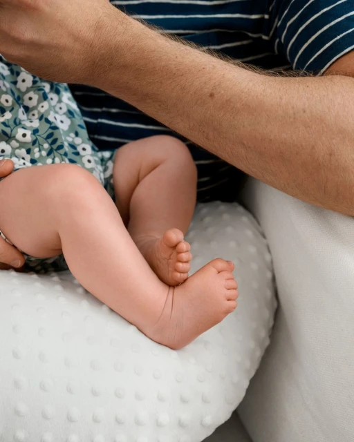 Close-up of a baby resting on a nursing pillow, with an adult's arm visible. The baby wears a floral print dress.