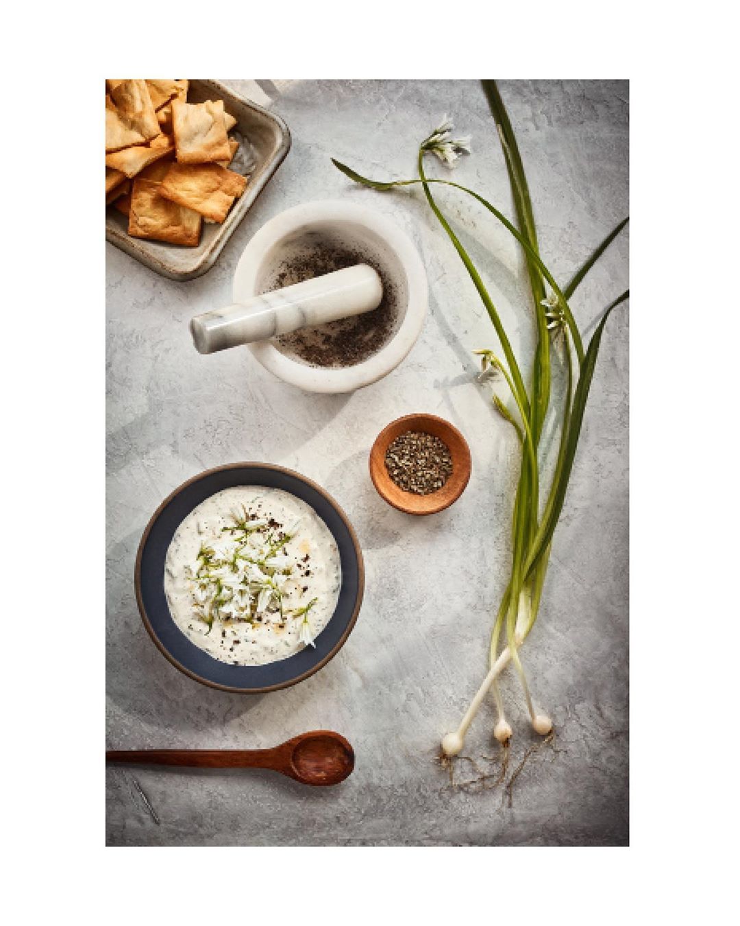 Overhead shot of a bowl of tzatziki sauce surrounded by crackers, a mortar and pestle, a wooden spoon, and a small bowl of spices.