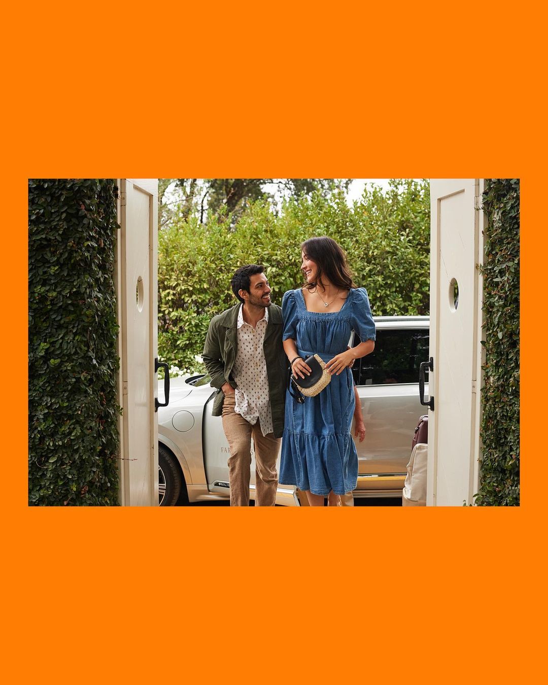 A stylish couple walks towards an open doorway on a sunny day; the woman wears a blue dress while the man wears a tan pants and green coat.