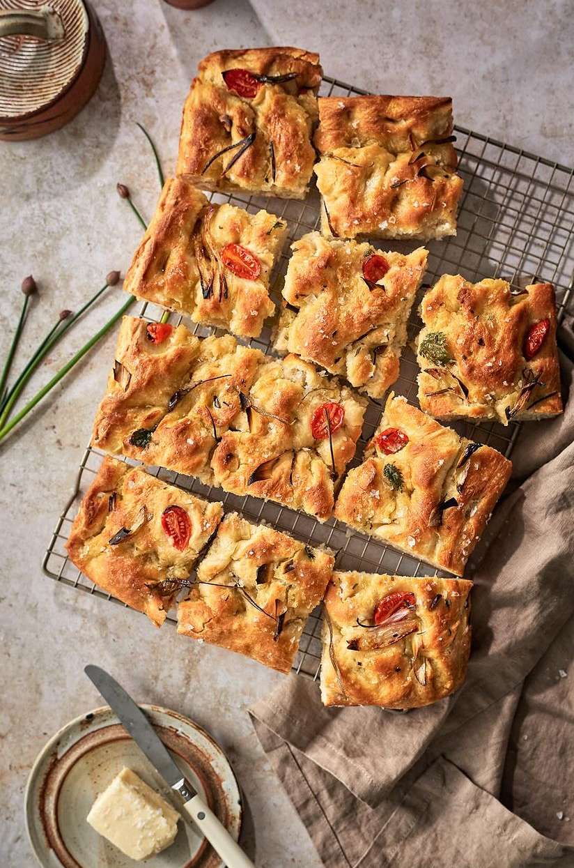 Overhead shot of tomato and onion focaccia bread squares arranged on a wire rack, with a knife and butter nearby on a table.