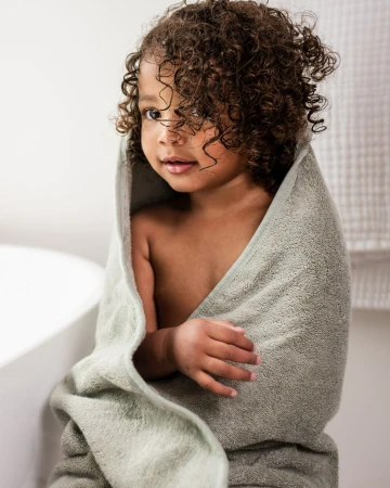 Adorable toddler with curly hair wrapped in a soft grey-green towel after bath time, standing in a bright, neutral bathroom.