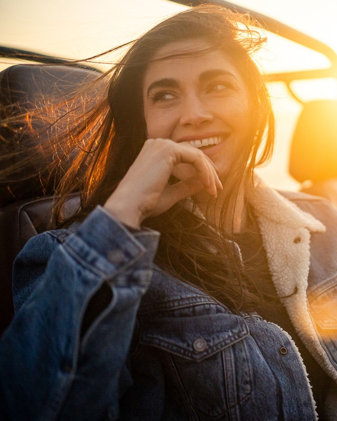 Smiling woman in a denim jacket rides in a car with bright sunlight in the background and her hair blowing in the wind.
