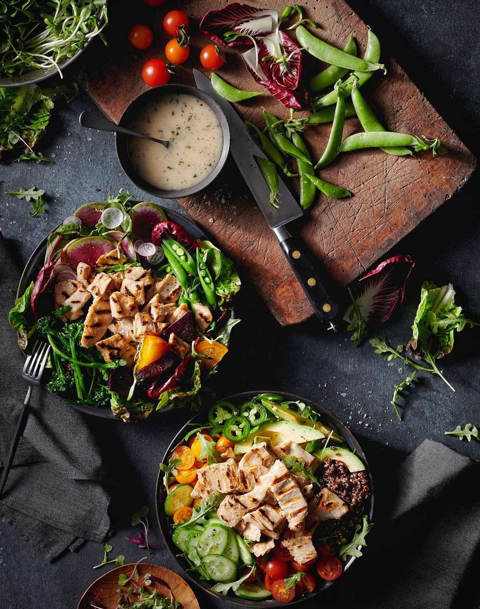 Overhead shot of two bowls with grilled chicken, vegetables, greens, salad dressing, and a knife on a wooden cutting board.