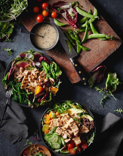 Overhead shot of two bowls with grilled chicken, vegetables, greens, salad dressing, and a knife on a wooden cutting board.