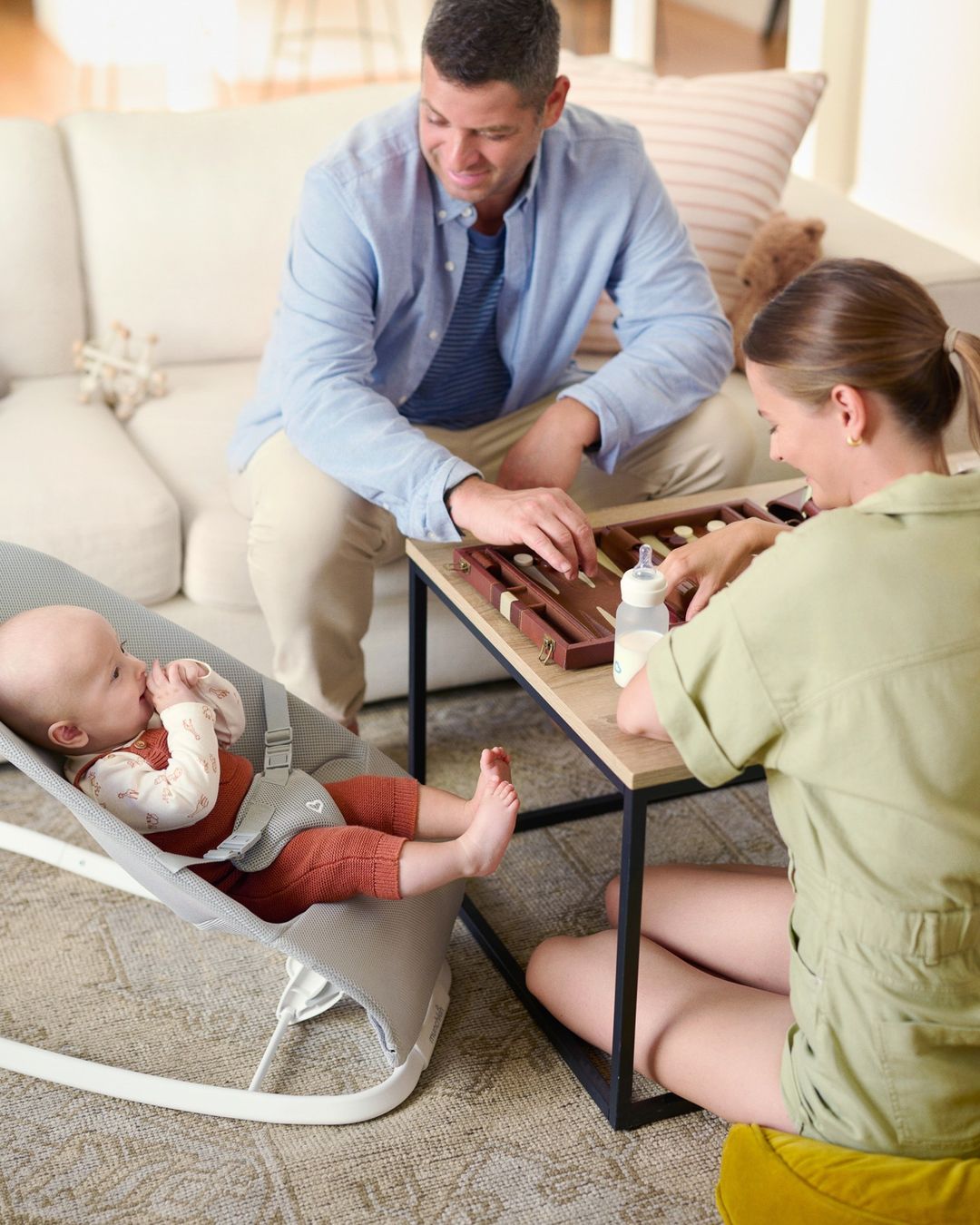 Parents play backgammon at a small table while their baby lies in a bouncer chair. A bottle sits on the table in the living room.