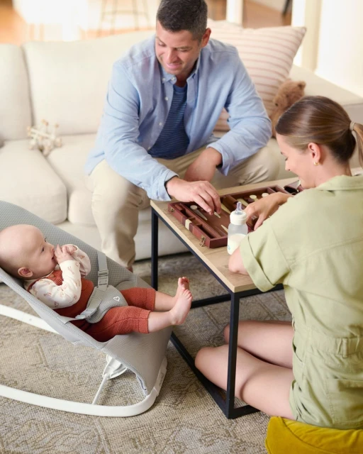 Parents play backgammon at a small table while their baby lies in a bouncer chair. A bottle sits on the table in the living room.