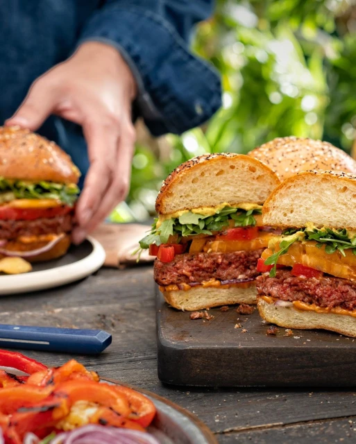 Vegan burgers served outdoors on a wooden table. Burgers include plant-based patties, lettuce, tomato, grilled peppers, and mustard.
