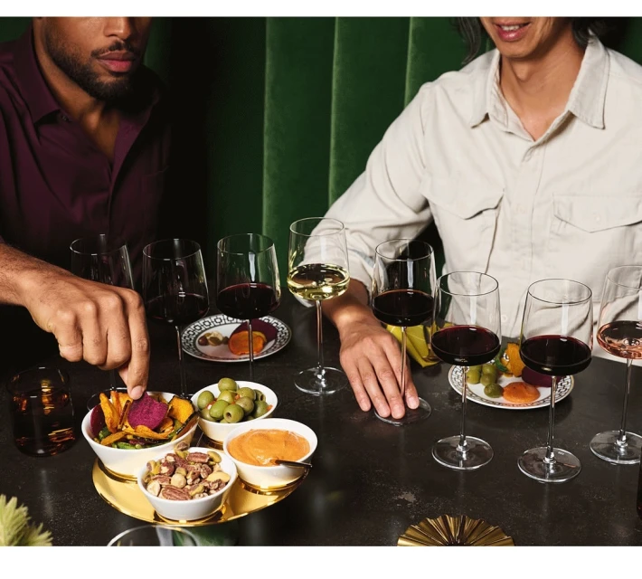 Two people at a wine tasting, with wine glasses and snacks arranged on a dark table against a green backdrop.