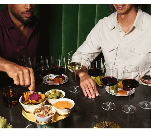 Two people at a wine tasting, with wine glasses and snacks arranged on a dark table against a green backdrop.
