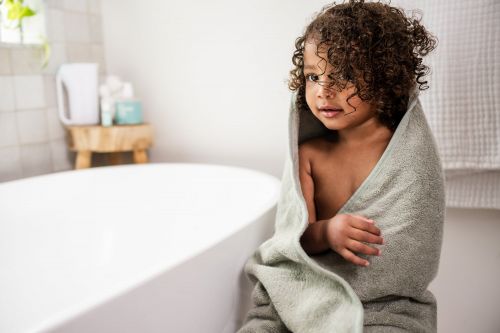 Child wrapped in a grey towel in a bright bathroom, looking off-camera with curly brown hair, next to a white bathtub.