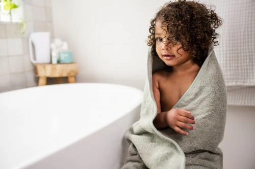 Child wrapped in a grey towel in a bright bathroom, looking off-camera with curly brown hair, next to a white bathtub.
