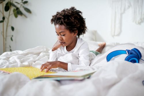 Boy reading book in bed with headphones, indoor daytime shot. Child engages with colorful book in a brightly lit bedroom.