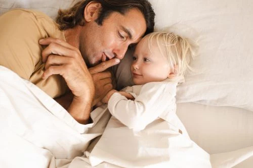 Father and toddler daughter bonding in bed, wrapped in white linens, sharing a tender moment with soft, natural lighting.