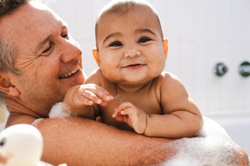 An adult holds a baby in a bubbly bathtub. Both are smiling and looking at the camera. A white rubber ducky sits near the side of the tub.