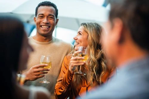 Two adults raise wine glasses in an outdoor social setting, laughing and smiling in the daytime with blurred figures in the foreground.