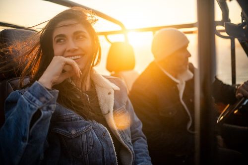 Young woman in a denim jacket smiles as she sits in a vehicle next to an older man, enjoying a sunlit ride.