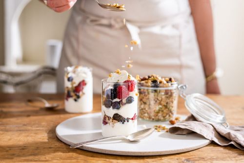 Granola being spooned over a layered parfait of yogurt, berries, and granola in a clear glass on a round white surface.
