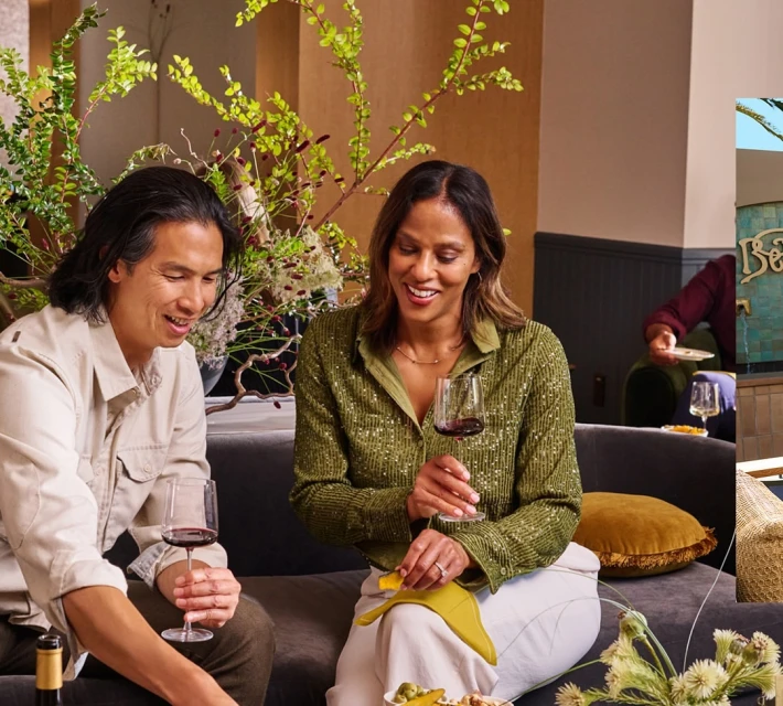 Two people, an adult male and female, sit with drinks and snacks in a lounge area with greenery and an outside view.