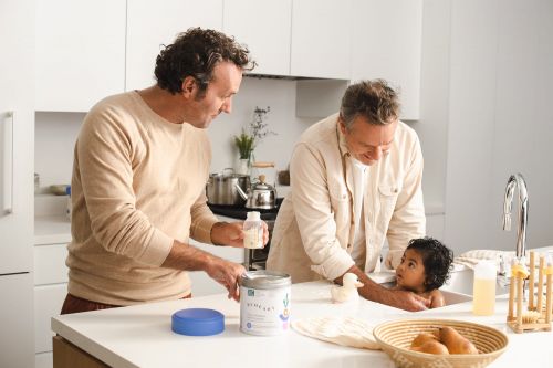 Two adult males give a baby a bath in the kitchen sink, one holding a bottle, showcasing a nurturing, modern family moment.