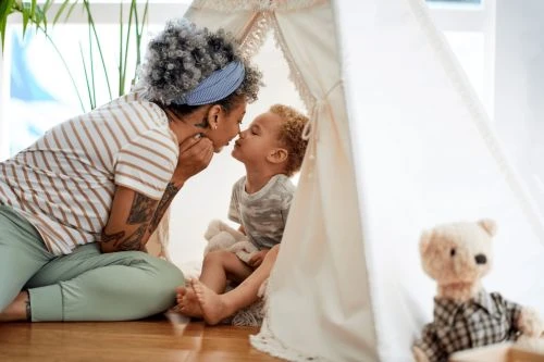 A mother and child share a tender moment inside a teepee tent indoors, with soft natural lighting and a teddy bear visible.