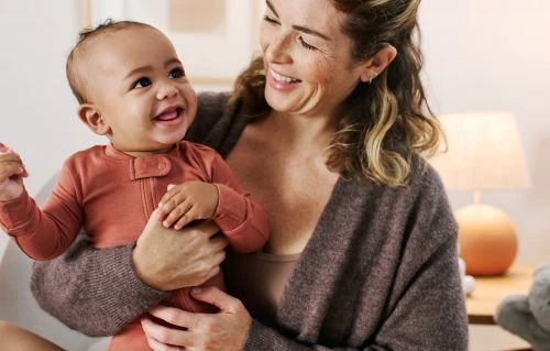 Smiling woman in a brown knit cardigan holding a happy baby in a terracotta romper indoors.