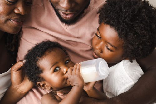 A close up of an African-American family with a baby drinking milk from a bottle, being held by an adult and child.