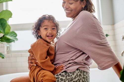 Smiling mother holds her young child in a bathroom, with a large green plant visible. The pair are laughing and looking at the camera.