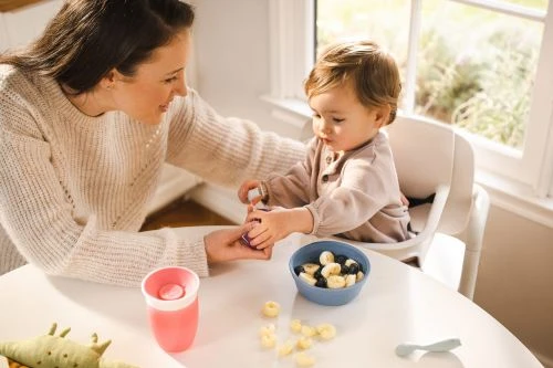 Young child in a high chair eating fruit and cereal from a bowl at a white table, assisted by an adult female in a cream sweater.