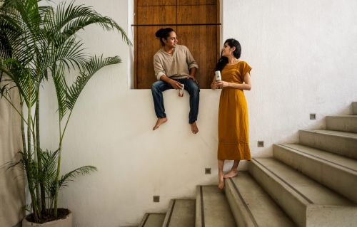 A young barefoot couple in casual clothing is sitting and standing on a staircase in a bright, white room near a potted palm tree.