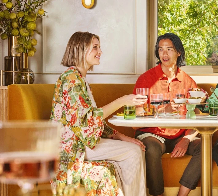 Woman in floral kimono and man in orange shirt sit at table with drinks in bright restaurant setting, enjoying a meal and conversation.