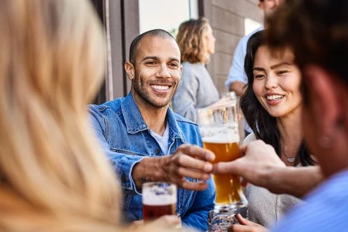 A group of friends at an outdoor bar smiles and toasts with glasses of beer in a cheerful, sunny setting, capturing a moment of camaraderie.
