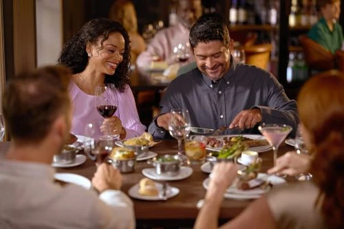Couple at dinner table in a restaurant, enjoying food and wine with friends, in a warmly lit, sophisticated indoor setting.