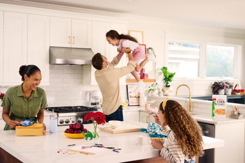 Family of four in a white kitchen. Father is holding up his young daughter while mother and older daughter are at the kitchen counter.