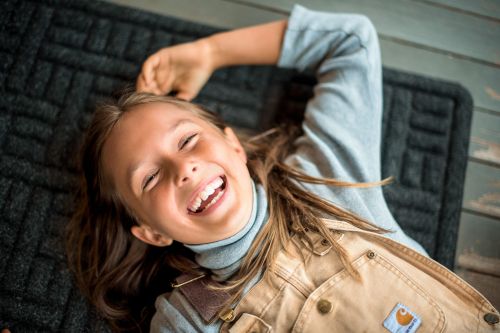 A young girl with long brown hair lies on a dark grey mat with a happy facial expression, wearing a blue turtleneck and tan overalls.