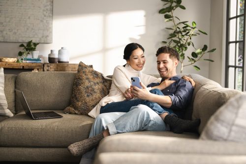 A smiling Asian woman and Caucasian man relax on a sofa looking at a smartphone in a well-lit indoor space with plants nearby.