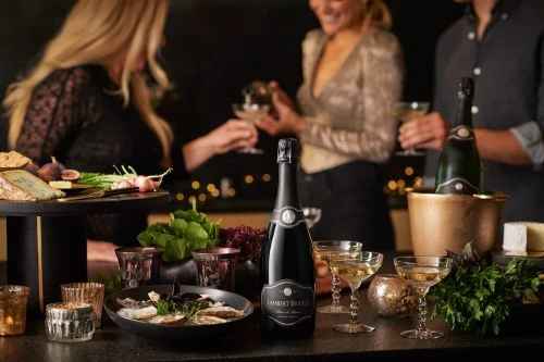 Three people celebrate with champagne glasses in an indoor setting. Champagne bottle in ice bucket with cheese, oyster, and radish appetizers.