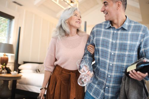 Elegant, mature couple with wine glasses and bottle, looking at each other in a hotel room with a bed and window in background.