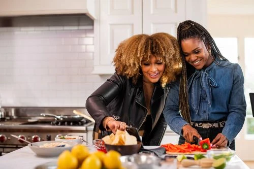 Two women are preparing food together in a kitchen, one cutting vegetables and the other looking at a phone with lemons and tortilla chips.
