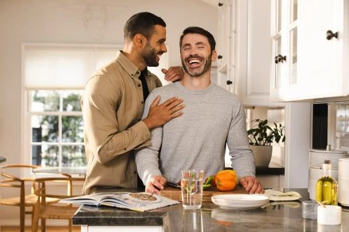 Two adult men in a kitchen: One touches the other's shoulder, both laughing near a countertop with cooking ingredients and a recipe book.