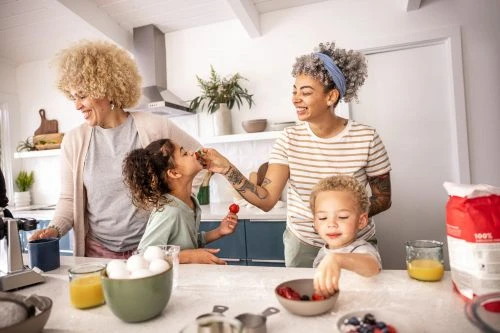 Two women and two children in a bright kitchen at a counter with food, enjoying a moment while preparing food and snacks together.
