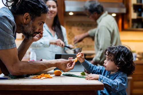 A father and child are at a counter with food, as two other adults prepare food in the blurred background in a home kitchen.