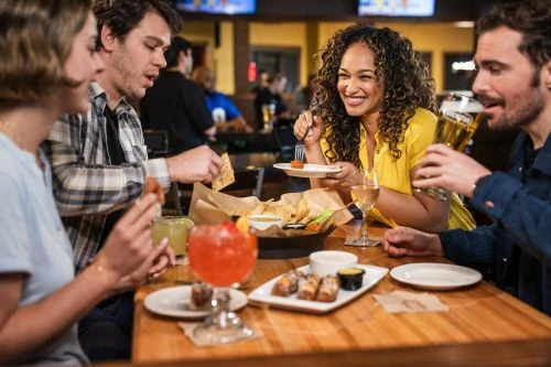 Group of four young adults dining at a restaurant with cocktails and appetizers on a wooden table, enjoying food and drinks together.