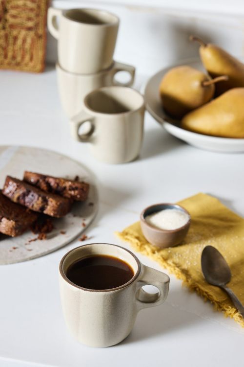 Still life on a white table featuring a cup of coffee, cake slices, sugar, pears in a bowl, a yellow napkin, and a spoon.