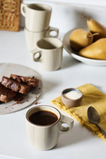 Still life on a white table featuring a cup of coffee, cake slices, sugar, pears in a bowl, a yellow napkin, and a spoon.
