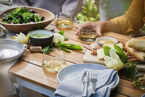 A rustic wooden table is set with dishes, silverware, glassware, bowls of food, and a salad. Two partially-visible people are seated at the table.