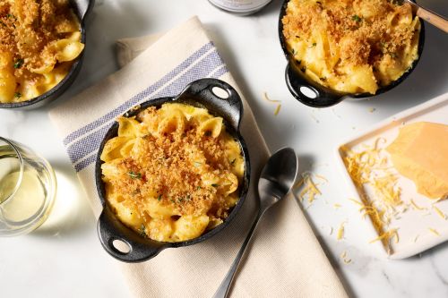 Overhead shot of macaroni and cheese in small cast iron pots with a spoon, cheese block and a glass of white wine on a white marble surface.
