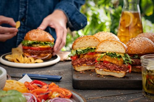Plant-based burger still life on a wood table, featuring an adult holding a chip, with leafy background.